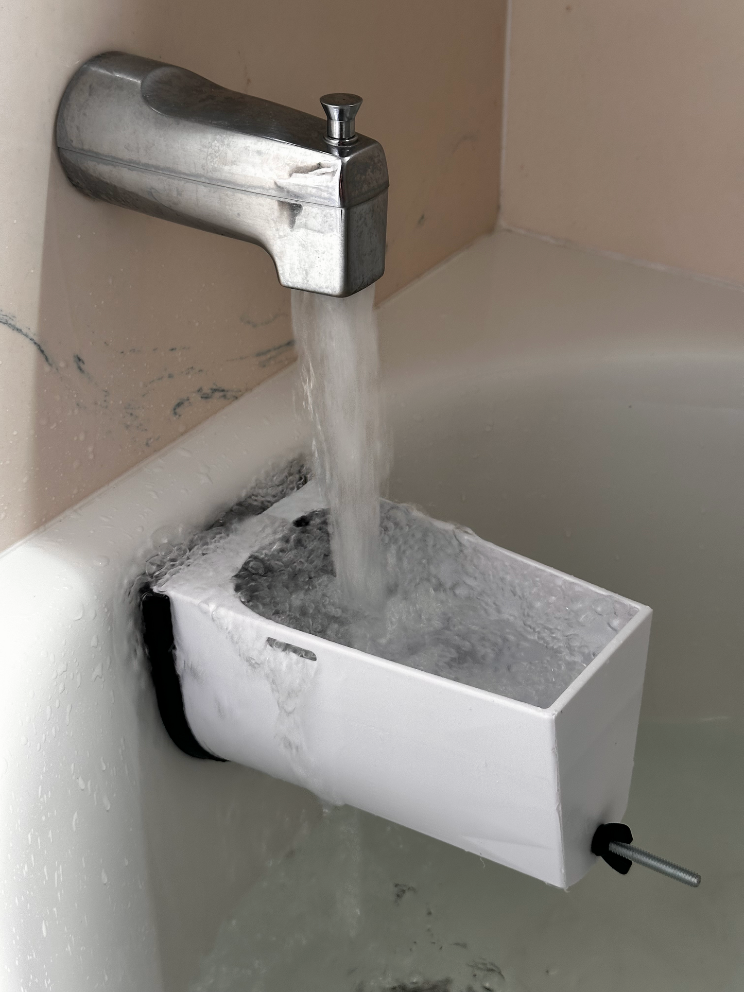 A white rectangular foam block with a black base and a metal rod is shown inside a bathtub, with water flowing from a chrome faucet into it.