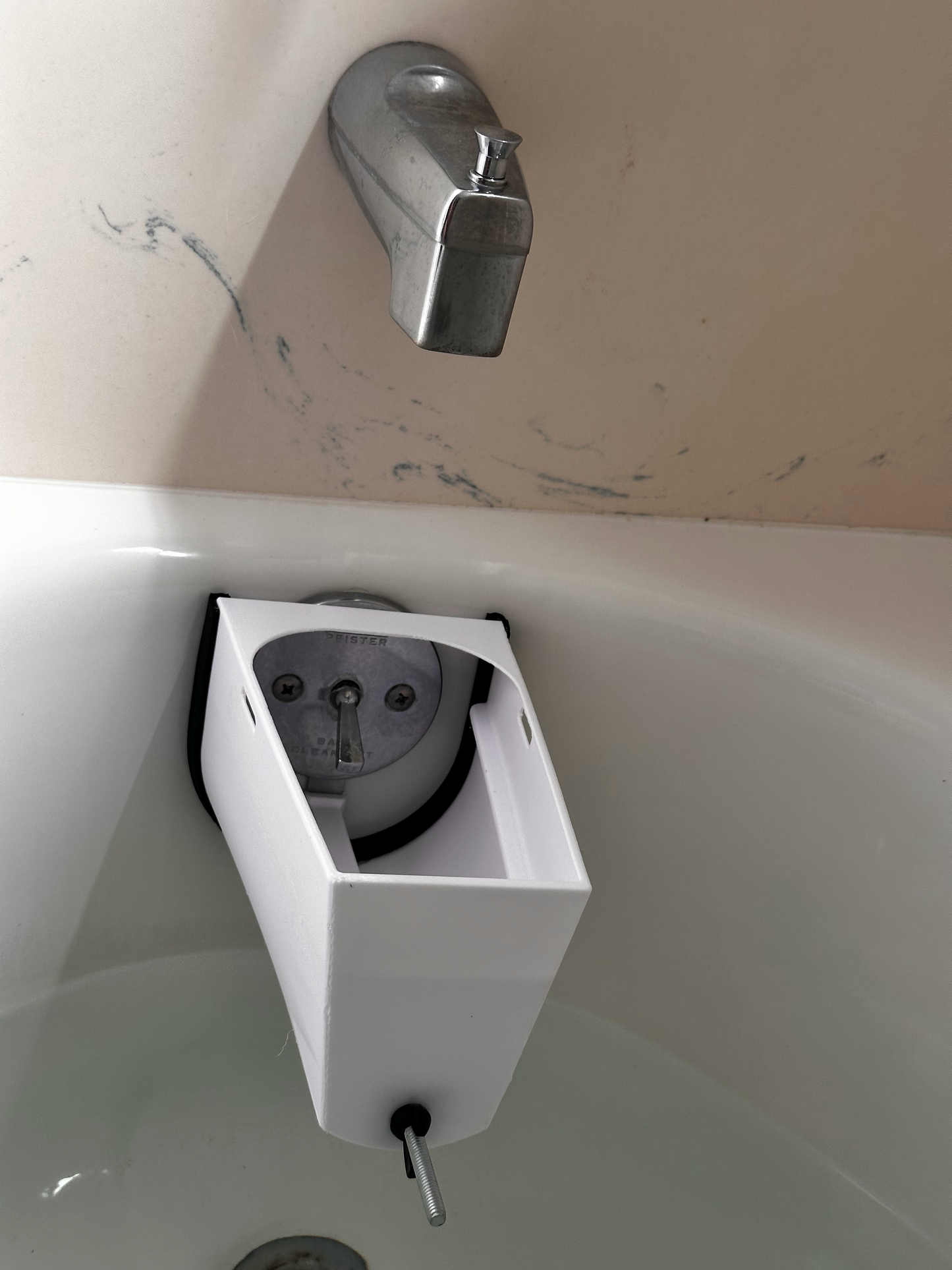 A white tub drain stopper or catchall mounted inside a tub basin, shown next to a chrome faucet on a beige wall.